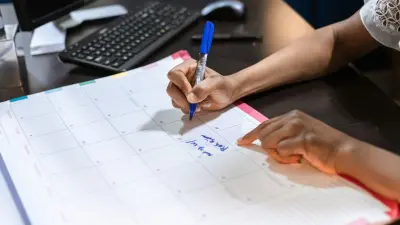 A hand writing a note on a paper desk calendar beside a keyboard