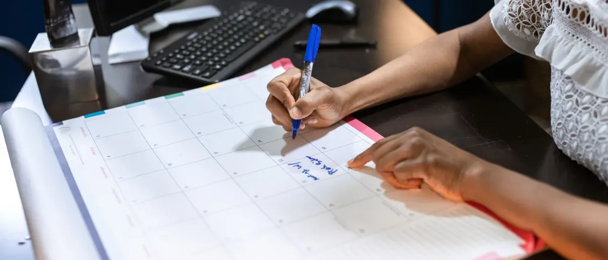 A hand writing a note on a paper desk calendar beside a keyboard