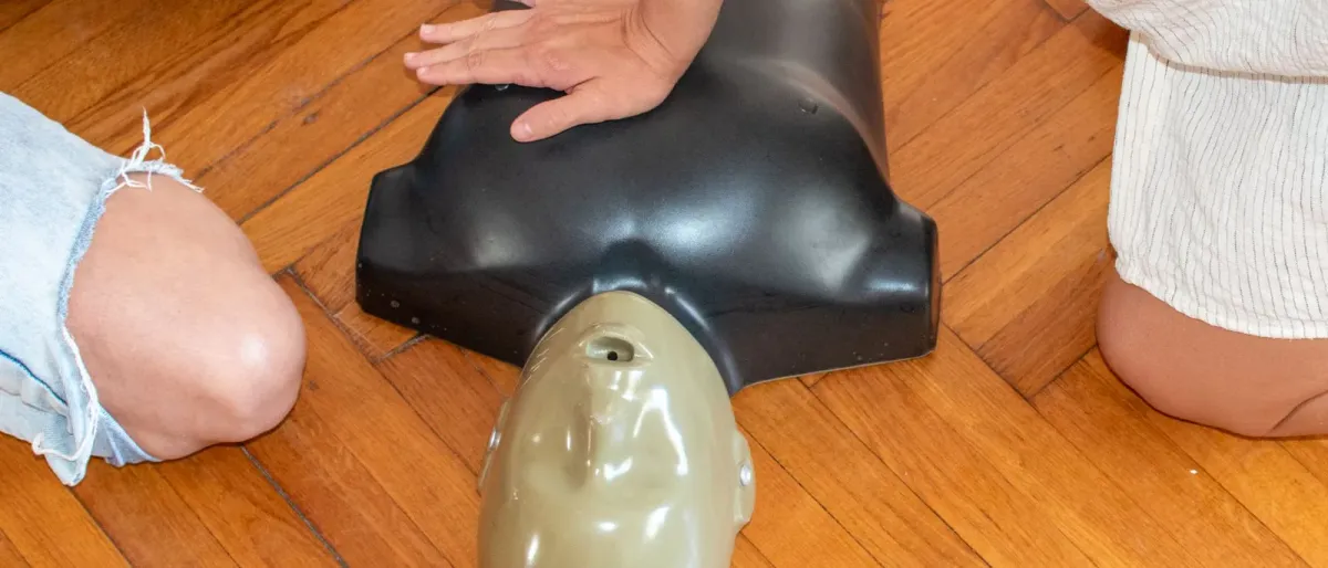 Hands practising chest compressions on a CPR training mannequin on a wooden floor