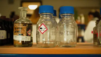 Laboratory reagent bottles on a shelf, each carrying a red-bordered hazard pictogram label