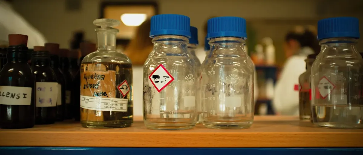 Laboratory reagent bottles on a shelf, each carrying a red-bordered hazard pictogram label