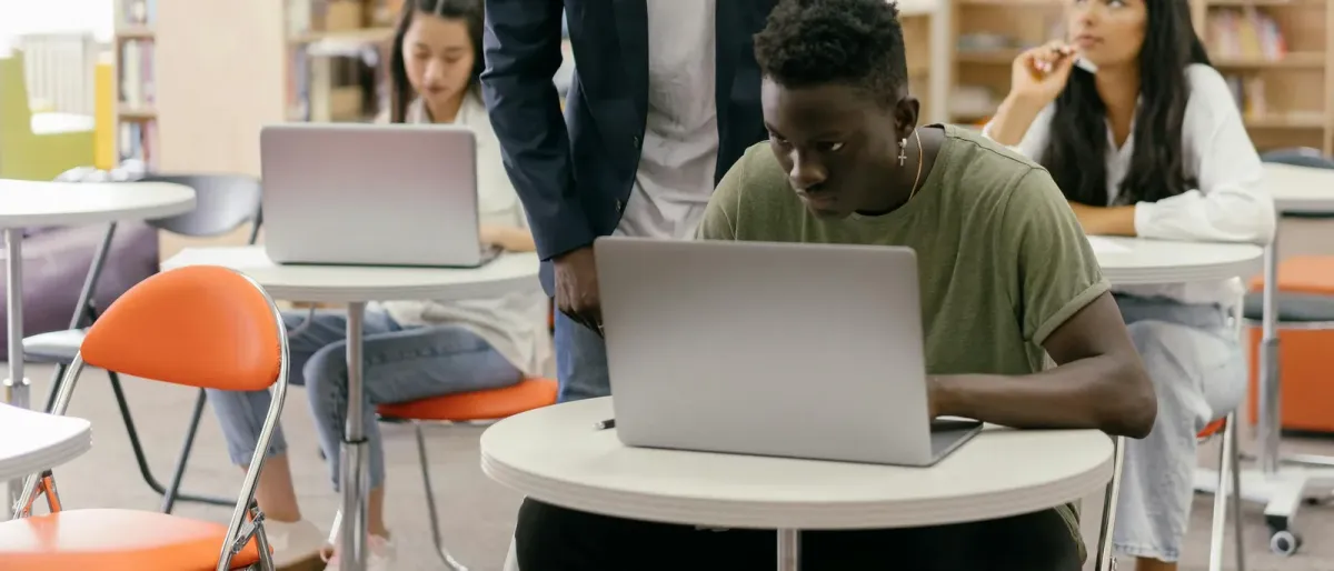Ontario high-school students working on laptops in a library-style ICT learning space while a teacher checks their progress