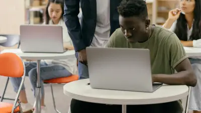 Ontario high-school students working on laptops in a library-style ICT learning space while a teacher checks their progress
