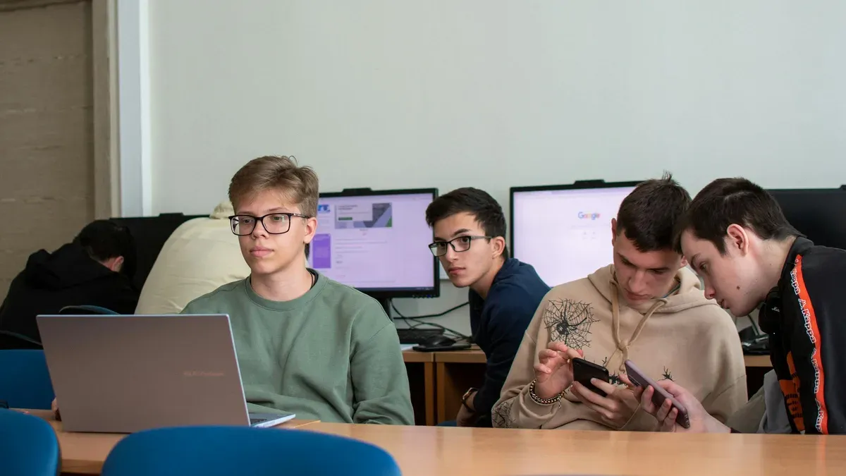 Grade 11 and 12 students collaborating at a high-school computer lab with laptops, desktop monitors, and mobile devices